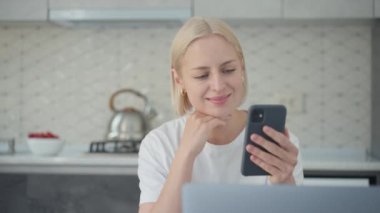 Cheerful smiling blonde woman actively listens and laughs during conference video call while sitting at home in kitchen.