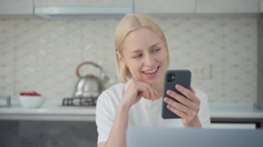 Young attractive smiling blonde woman talks and gesticulates in video conference call while sitting at home in kitchen.
