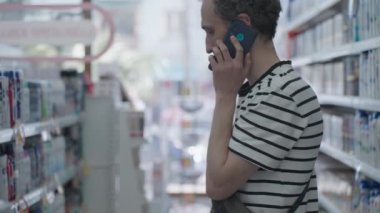 Young handsome curly brunet talks on phone between shelves with hygiene products. Adult man in striped black and white t-shirt inside in health and beauty department. High quality 4k footage