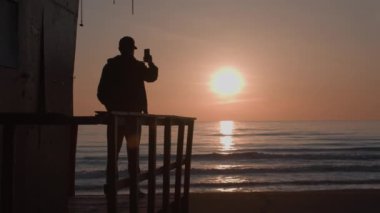 Silhouette of young man on observation deck shooting seaside sunset on phone. Yound adult male tourist makes a photo of sundown on clear sky, calm sea and empty beach. Slow motion. High quality 4k