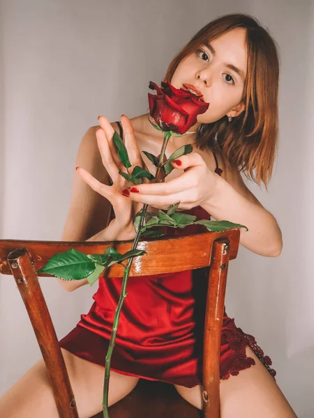 赤い無視者の古い木製の椅子の上に魅力的な細い若い赤髪の女性は花弁の上に水滴と大きな赤いバラの香りを楽しんでいます — ストック写真