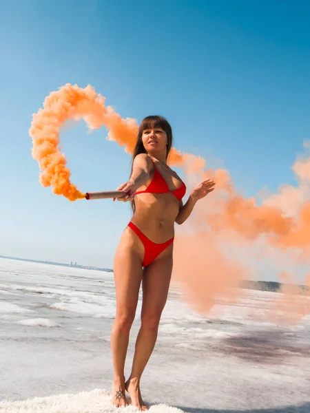 赤いビキニの水着を着た長い暗い髪の女性が トーチ花火でほっそりと日焼けした体でピンクの煙の人形を空中に描きます アクティブな健康的なライフスタイル フィットネス アウトドアスポーツ 極端なレクリエーションの概念 — ストック写真