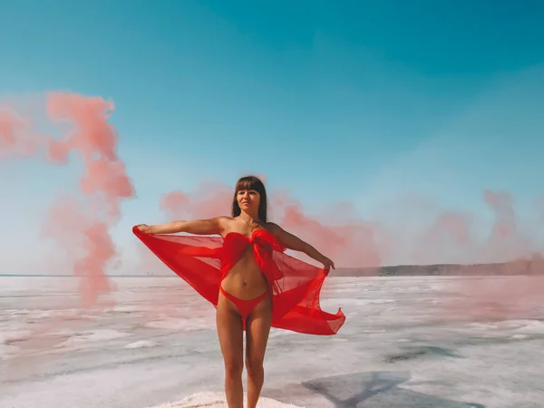 赤いビキニの水着で魅力的な運動体と赤い煙で氷の上で踊るシルクの赤いケープを持つ美しい女性は アクティブなライフスタイル 屋外レクリエーション 極端なレクリエーション 冬のレクリエーション ダンスクラス 花火の概念 — ストック写真