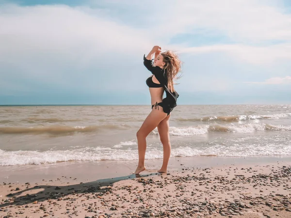 夏日里 在海浪的映衬下 一个身穿黑色开放衬衫和黑色比基尼泳衣 身材优美的金发女子在沙滩上跳舞 — 图库照片