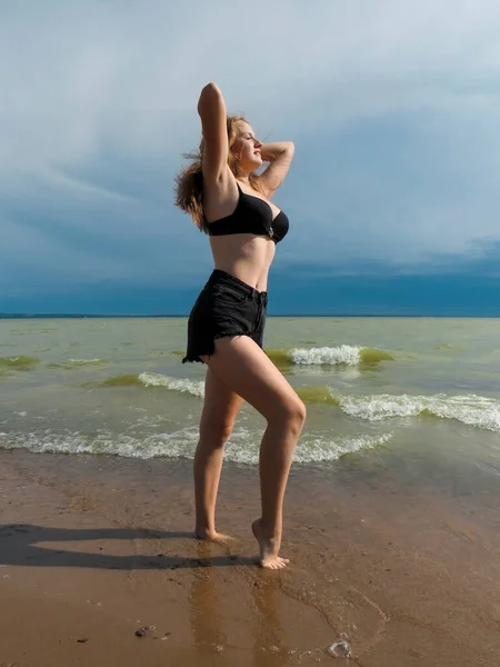 一位身穿黑色短裤和黑色胸罩的金发女子 身材苗条 身材苗条 身披黑色胸罩 在沙滩上跳舞 背景是海浪 在暴风雨即将来临的夏日 蓝蓝的天空刮起了大风 — 图库照片