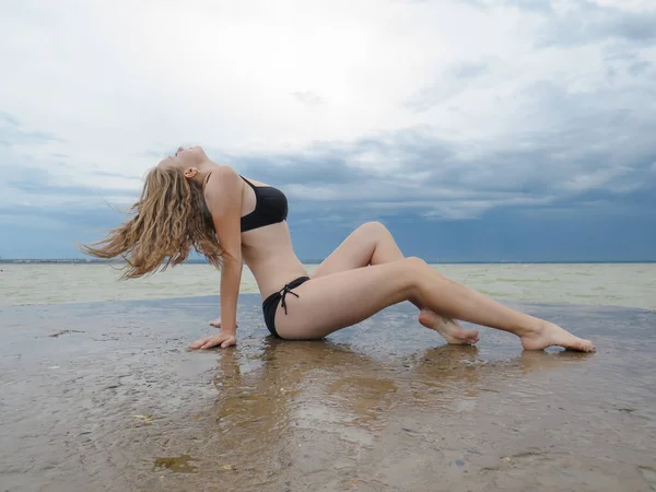 年轻的性感女子 身穿黑色比基尼 体形苗条 金发飘逸在码头的湿石板上 背景是夏日风暴前的水雾和乌云 — 图库照片