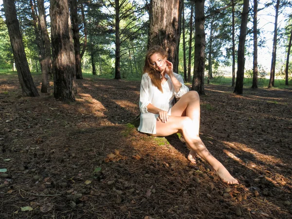 一个赤脚的年轻女子 一头长长的金发 光着细长的腿 穿着白色的丝质衬衫 站在树干旁 站在针叶林里 夏日阳光灿烂 蓝天映衬下 美丽与青春的概念 健康的生活方式 — 图库照片