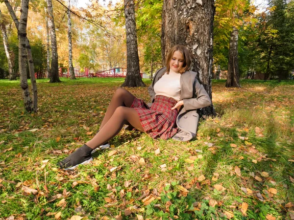 年轻的金发女子穿着漂亮的长腿紧身衣 灰色外套和短短的格子裙 站在城市公园一棵大树的树干旁 在阳光温暖的九月里 金秋的概念 自然漫步 — 图库照片