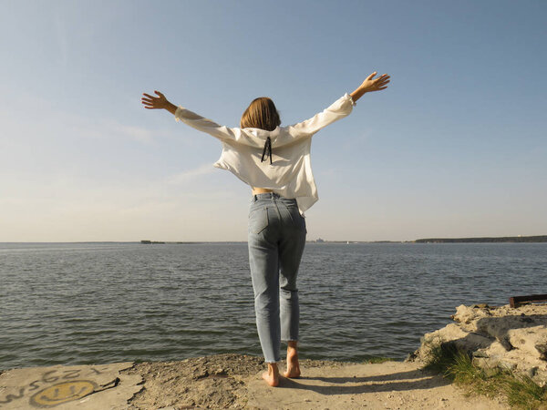 A barefoot blonde young woman in a white silk shirt and blue denim pants on an old stone pier against the background of the water of the sea bay enjoys a rest in nature on a clear sunny day