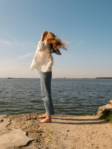 A barefoot blonde young woman in a white silk shirt and blue denim pants on an old stone pier against the background of the water of the sea bay enjoys a rest in nature on a clear sunny day