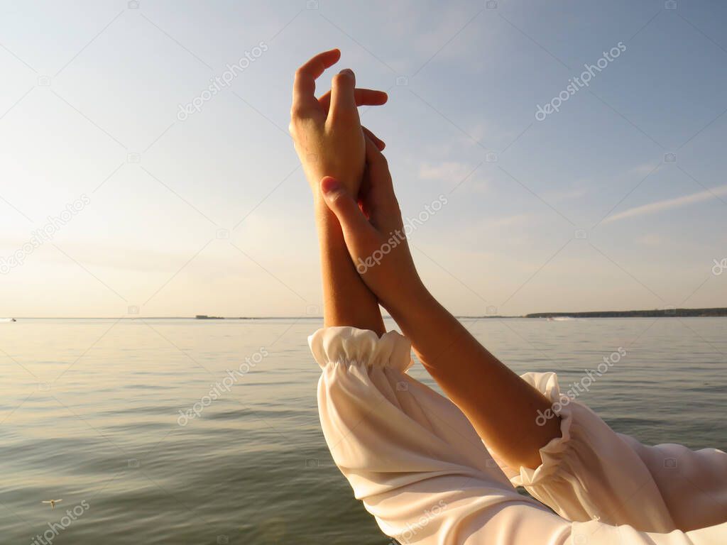 Beautiful hands of a young woman on the background of the water surface of the sea in the rays of the sun at sunset, close-up, the concept of harmony and unity with nature, rest on the beach