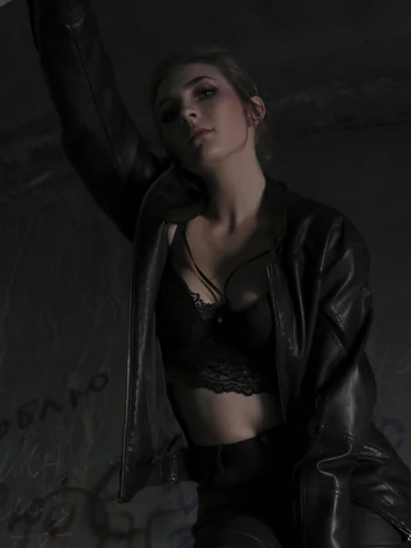 一个年轻貌美的女人 身材苗条 身穿黑色皮夹克和紧身裤 头戴黑色胸罩 靠着一栋废弃建筑的砖墙 — 图库照片