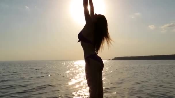 夏の夜の夕日の海のビーチの水の背景に太陽のオレンジ色の光の中でビキニ水着で若い魅力的な女性 美しさと健康の概念 ビーチの休日 日焼け 屋外休暇 — ストック動画