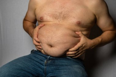 Şişman, üstsüz, beyaz bir adam oturuyor ve yağlarını tutuyor.