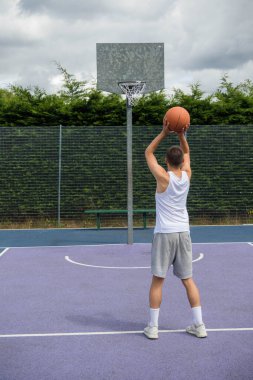 19 yaşında bir ergen çocuk halka açık bir parkta basketbol sahasında şut çekiyor.