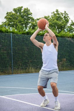 19 yaşında bir ergen çocuk halka açık bir parkta basketbol sahasında şut çekiyor.