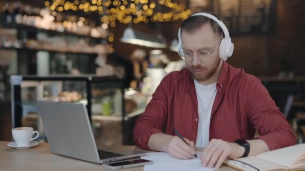 Klidný soustředěný kavkazský student nebo pracovník v brýlích se při posezení v kavárně dívá na online přednášky nebo webináře a píše poznámky do sešitu. Koncept technologií. — Stock video