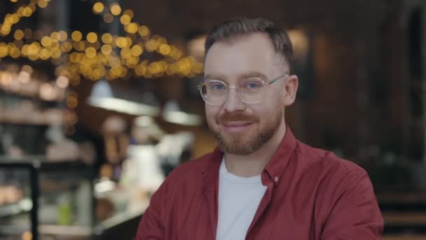 Portrétní pohled na veselého vousatého studenta s brýlemi na kameře sedícího v kavárně. Obchodník v neformálním oblečení pózuje na kameru, zatímco sedí u stolu. — Stock video