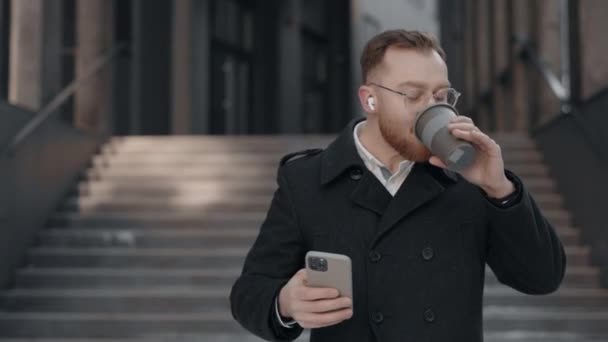 Akıllı telefon kullanıp kahve içen yakışıklı bir işadamı iş merkezinin yakınında yürüyor. Genç adam dışarıda cep telefonunu karıştırıyor. İş için. Sosyal ağ. Uygulama. — Stok video