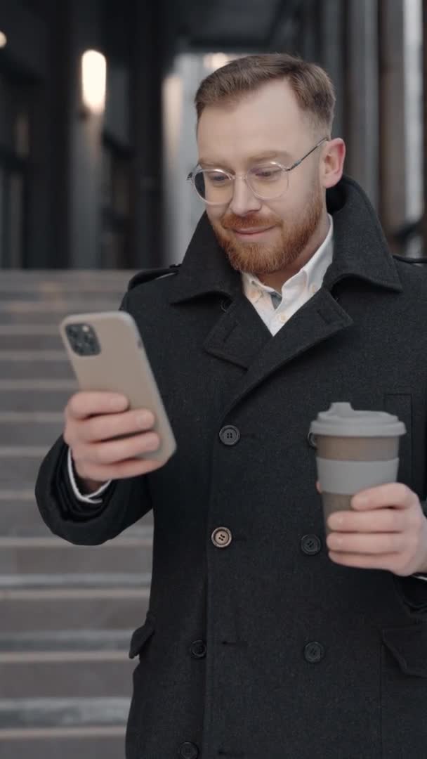 Verticale video van de zakenman in formele pak met behulp van smartphone en het vasthouden van kopje koffie in handen staan in de buurt van business center. Communicatie. Apps. — Stockvideo