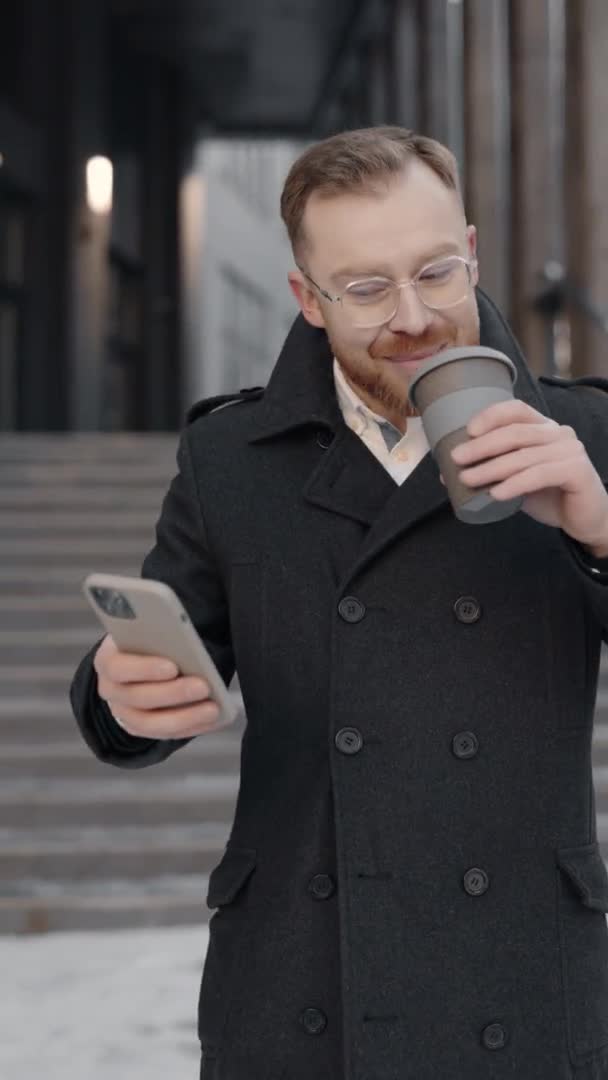 オフィスの近くを歩くスマートフォンを使用して正式なスーツのビジネスマンの垂直ビデオ。スマートフォンで白人男性のテキストメッセージとコーヒー屋外のカップを保持.コミュニケーション. — ストック動画