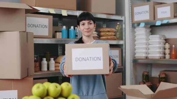 Egy fehér nő portréja, aki kartondobozt tart élelmiszerrel, miközben jótékonysági alapnál áll. Női barna önkéntes munka az élelmiszerbankban a rászorulók megsegítésére. — Stock videók