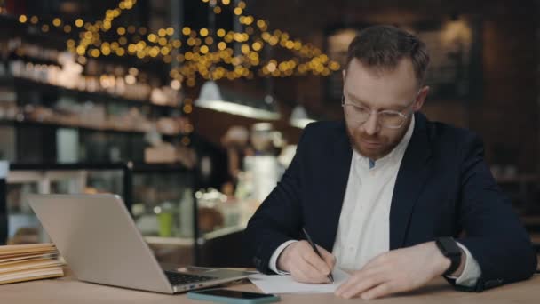 Επιχειρηματίας κρατώντας σημειώσεις, ενώ κάθεται στο τραπέζι με το φορητό υπολογιστή στο καφέ. — Αρχείο Βίντεο