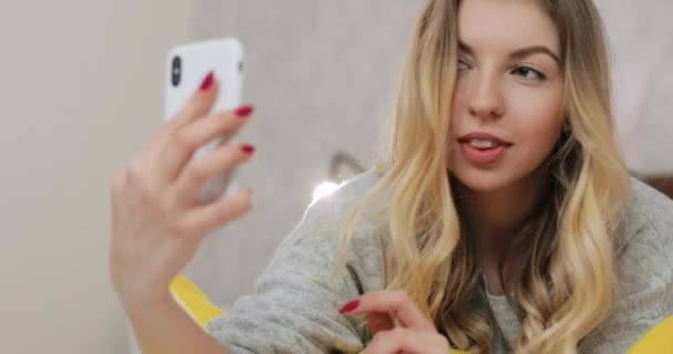 Giovane donna caucasica sorridente a letto scattando selfie foto e video con fotocamera frontale dello smartphone. Stile di vita e concetto di tecnologie. — Video Stock