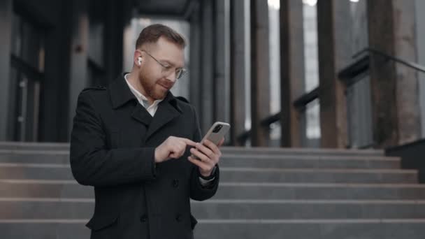 Biały biznesmen w garniturze używa smartfona stojącego w pobliżu biurowca. mężczyzna pracodawca stukanie lub smsowanie na jego smartfon. — Wideo stockowe
