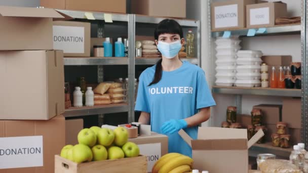 Női önkéntes arcmaszkban és kesztyűben pózol az élelmiszerbankban — Stock videók