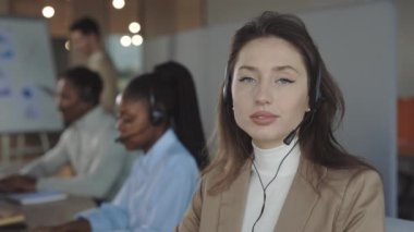 Çağrı merkezinde çalışırken gülümseyen ve kameraya bakan mutlu beyaz kadın portresi. Çok ırklı çalışan müşteri hizmetleri temsilcileri arka planda telefonla konuşuyor..