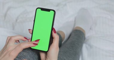 LVIV, UKRAINE - 5 Mart 2022: Evdeki yatakta yeşil ekranlı akıllı telefon kullanan genç kadın. Beyaz kadın, elinde modern aletlerle yatağında yatıyor. Kapat..