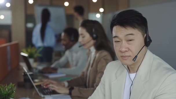 Retrato de hombre asiático feliz sonriendo y mirando a la cámara mientras trabajaba en el centro de llamadas. Grupo multiétnico de representantes de servicio al cliente de los trabajadores hablando por teléfono en el fondo. — Vídeos de Stock