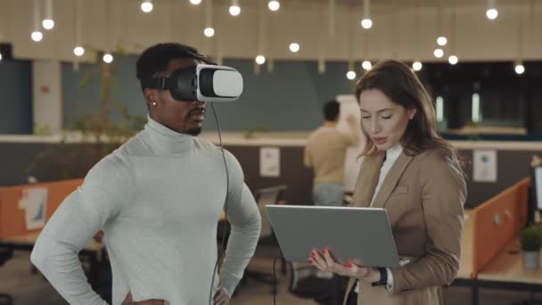 Un homme d'affaires afro-américain teste un casque simulateur de réalité virtuelle et fait des gestes en l'air pendant que sa collègue analyse les gestes et enregistre sur un ordinateur portable. Personnes travaillant au bureau. — Video