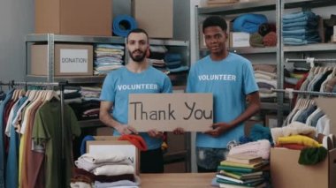 Dolap afişi taşıyan çeşitli erkek gönüllülerin portre görüntüsü teşekkür cümlesiyle ve gülümseyerek kameraya bakıyor. Arkasında eşyalar olan raflar. İnsani yardım kavramı.