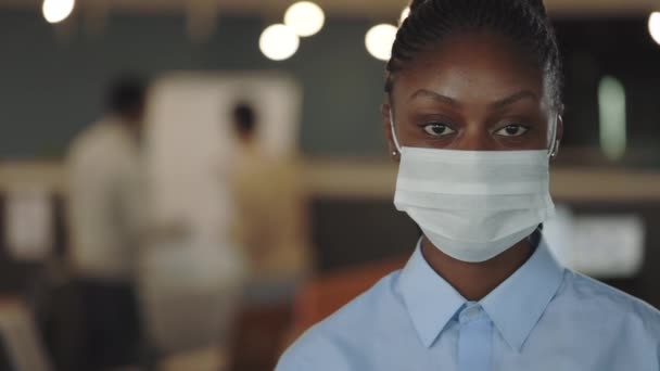 Tıbbi maske takmış genç Afro-Amerikan iş kadını modern ofiste duran kameraya bakıyor. Ofis odasındaki bir adamın portresi. Coronavirus konsepti. — Stok video