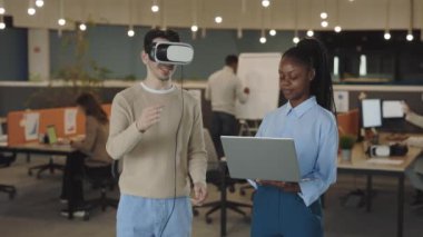 Beyaz iş adamı, sanal gerçeklik simülatörü kulaklığını test ediyor ve kadın meslektaşı el hareketlerini analiz edip bir dizüstü bilgisayarda kayıt yaparken havada el kol hareketi yapıyor. Ofiste çalışanlar.