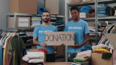 Bağış sözcüğüyle birlikte dolaptan pankart tutan ve kameraya gülümseyerek bakan çeşitli gönüllülerin portre görüntüsü. Arkasında eşyalar olan raflar. İnsani yardım kavramı.