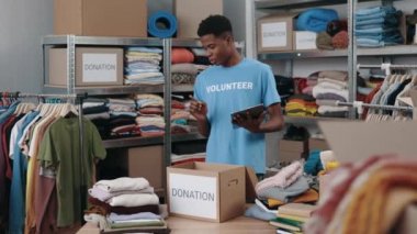 Esmer adam tabletin başında yazarken depoda duruyor ve bağış için kıyafet almayı düşünüyor. Arka plandaki raflara aitti. İnsani yardım kavramı.