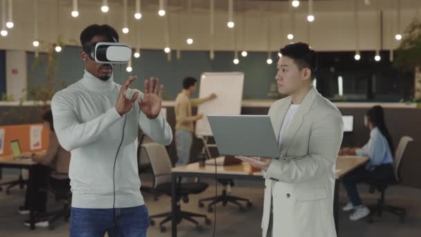 Un homme d'affaires afro-américain teste un casque simulateur de réalité virtuelle et fait des gestes en l'air pendant que son collègue masculin analyse les gestes et enregistre sur un ordinateur portable. Personnes travaillant au bureau. — Video