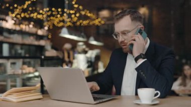 Kafede çalışan bir iş adamı, dizüstü bilgisayarla oturuyor, akıllı telefonu tutuyor, konuşuyor..