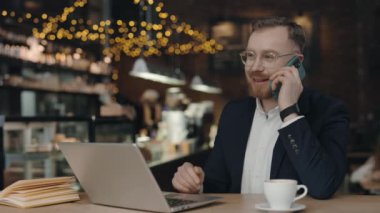 İş adamı kafeteryada oturuyor, akıllı telefonla konuşuyor ve gülümsüyor, müşterilerle ya da iş arkadaşlarıyla telefonda konuşuyor. Uzak çalışma.