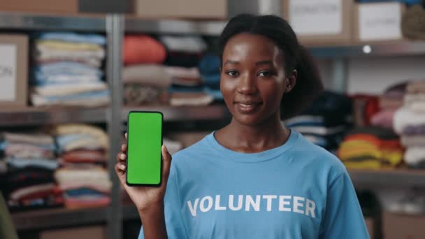 Widok na portret wielorasowej kobiety trzymającej smartfona z zielonym, makietowym ekranem i pokazującej go przed kamerą stojąc w centrum darowizny. Koncepcja wolontariuszy. — Wideo stockowe