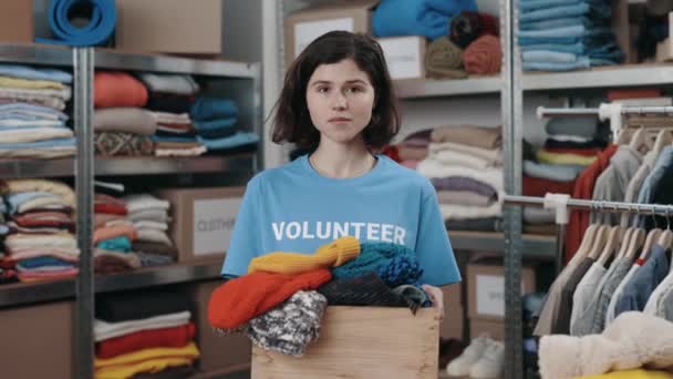 Portret van de blanke vrijwilliger vrouw die doos met kleren vasthoudt en glimlachend naar de camera kijkt. Planken met bezittingen op de achtergrond. Donatieconcept. — Stockvideo