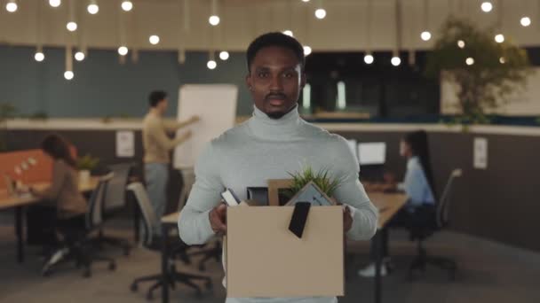 Portret bezrobotnego, afrykańskiego mężczyzny, zwolnionego z pracy. Zdenerwowany młody człowiek z pudełkiem pełnym rzeczy w rękach patrzących przed kamerę. W środku. Strzelam z pracy. Kryzys i biznes. — Wideo stockowe