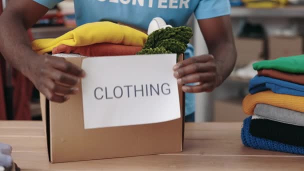 Voluntário multirracial colocando papel com palavra de vestuário na caixa do armário com roupas de doação no armazém. Ajuda humanitária e conceito de doação. Vista cortada. — Vídeo de Stock