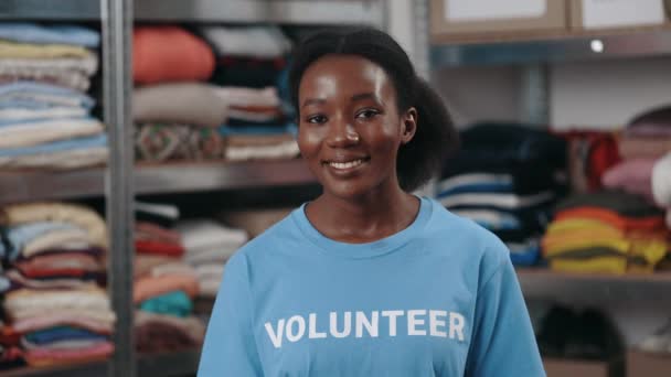 Πορτρέτο άποψη της πολυφυλετικής γυναίκας σε εθελοντική t πουκάμισο κοιτάζοντας την κάμερα με χαμόγελο. Ράφια με αντικείμενα για δωρεά στο παρασκήνιο. Ανθρωπιστική βοήθεια. — Αρχείο Βίντεο