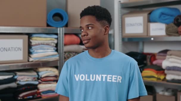 Porträtt bild av multiracial man i volontär t skjorta tittar på kameran med leende. Hyllor med tillhörigheter för att donera i bakgrunden. Begreppet humanitärt bistånd. — Stockvideo