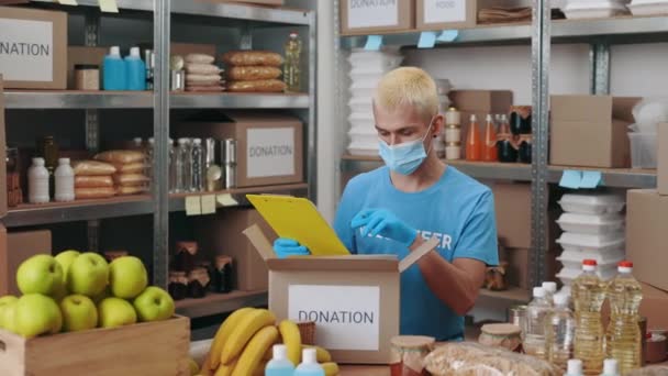Człowiek w masce piszący na schowku wśród banków żywności — Wideo stockowe