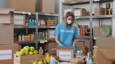 Yüz maskeli ve eldivenli kadın çantaları bağışlanmış yiyeceklerle dolduruyor.